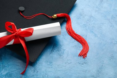 Diploması kırmızı kurdeleli ve mezuniyet şapkası mavi masada.