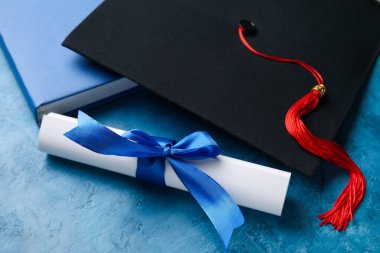 Kurdeleli diploma, mezuniyet şapkası ve mavi masadaki kitap.