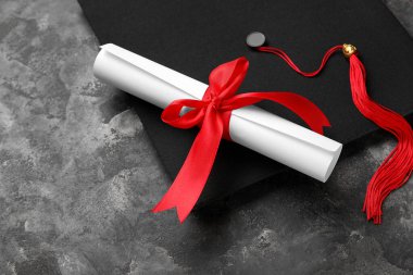 Kırmızı kurdeleli diploma ve koyu gri masada mezuniyet şapkası.