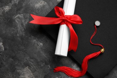 Kırmızı kurdeleli diploma ve koyu gri masada mezuniyet şapkası.