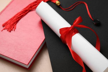 Kırmızı kurdeleli diploma, mezuniyet şapkası ve bej masa üzerine kitap.