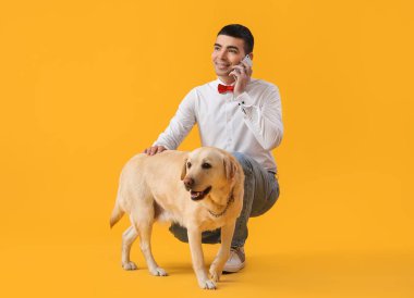 Labrador köpeği olan genç adam sarı arka planda cep telefonuyla konuşuyor.
