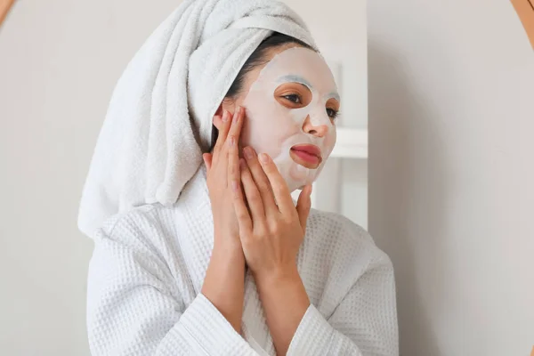 Young woman with sheet mask near mirror in bathroom, closeup