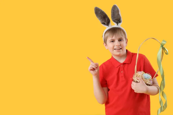 黄色の背景に何かを指してイースターの卵のバスケットを持つ小さな男の子 — ストック写真