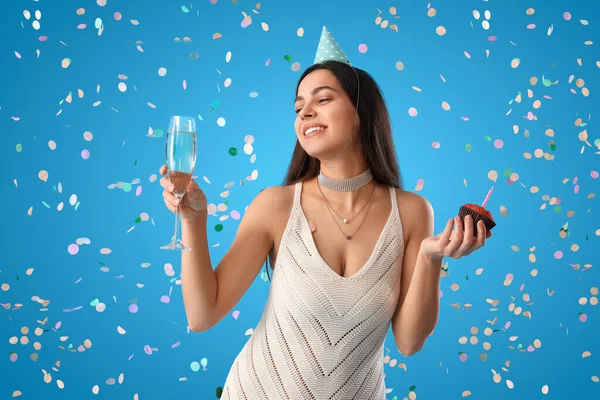 Happy young woman with Birthday muffin and glass of champagne on blue background