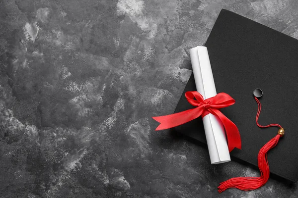 stock image Diploma with red ribbon and graduation hat on dark grey table