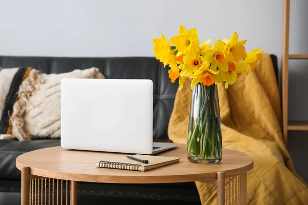 Modern laptop, vase with narcissus flowers and notebook on coffee table in living room