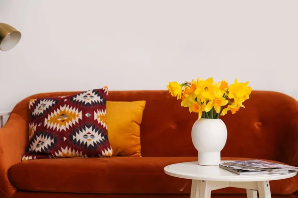 Vase with narcissus flowers and magazines near sofa in living room