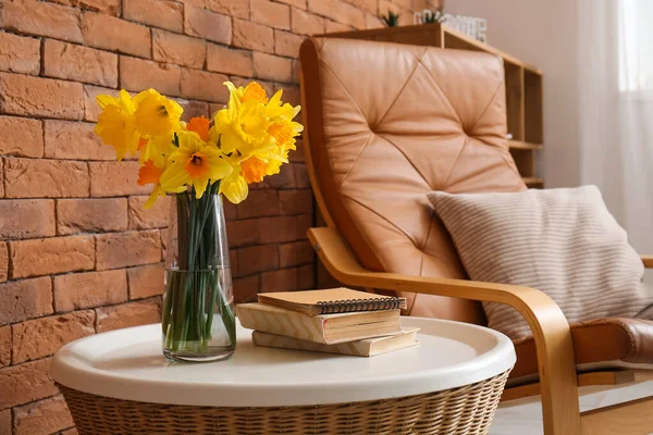 Vase with narcissus flowers and books on coffee table near armchair in living room