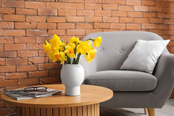 Vase with narcissus flowers and magazines on coffee table near cozy armchair in living room