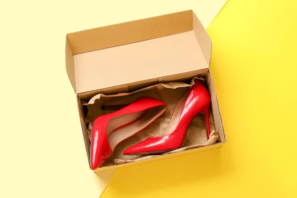 stock image Cardboard box with high heeled shoes on color background