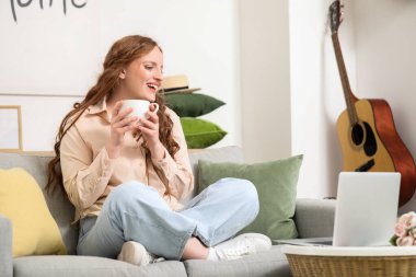 Evdeki kanepede oturan bir fincan çayla güzel kızıl saçlı kadın.