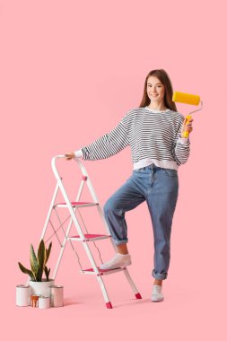 Young woman with roller, ladder, paint cans and houseplant on pink background