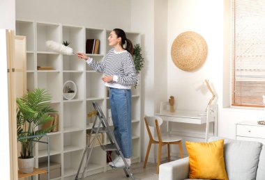 Young woman on stepladder cleaning bookshelf with duster at home