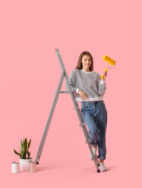 Young woman with roller, ladder, paint cans and houseplant on pink background