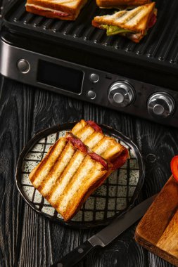 Siyah ahşap masada, modern elektrikli ızgaranın yanında lezzetli sandviçler.