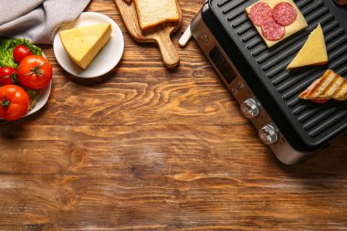 Modern elektrikli ızgara, lezzetli sandviçler ve ahşap arka planda malzemeler.