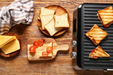 Ahşap arka planda lezzetli sandviçler ve malzemelerle modern elektrikli ızgara.