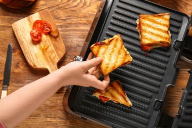 Ahşap arka planda modern elektrikli ızgaranın yanında nefis bir sandviç tutan kadın.