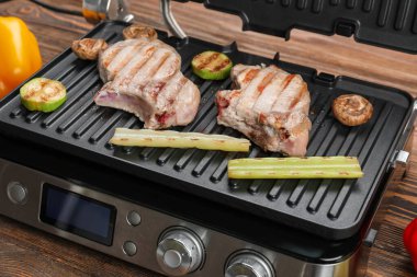 Ahşap arka planda biftek ve sebzeli modern elektrikli ızgara.