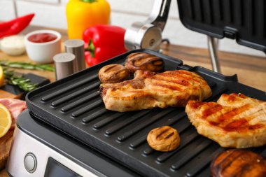 Hafif tuğla duvarın yanındaki masada lezzetli biftekleri ve mantarları olan modern elektrikli ızgara, yakın plan.