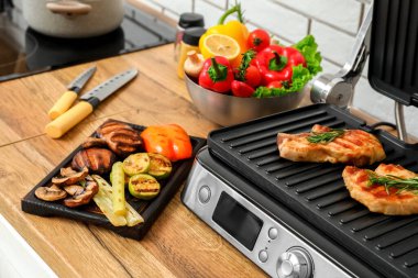 Hafif tuğla duvarın yanındaki masada lezzetli biftek ve sebzeli modern elektrikli ızgara, yakın plan.