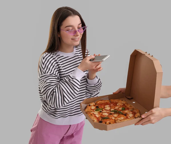 Young woman with box of tasty pizza using mobile phone on grey background