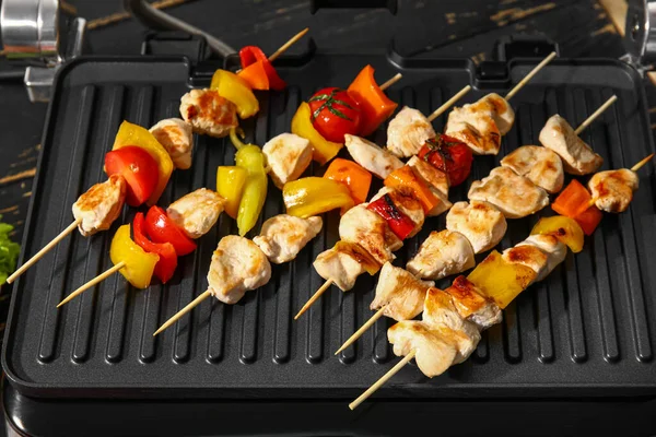 Modern electric grill with tasty chicken skewers and vegetables on dark wooden background