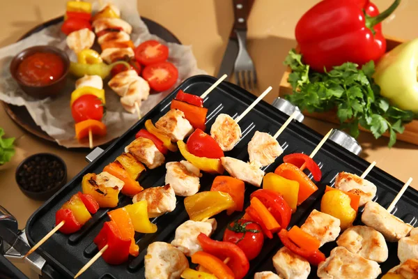 Modern electric grill with tasty chicken skewers and vegetables on beige wooden background, closeup