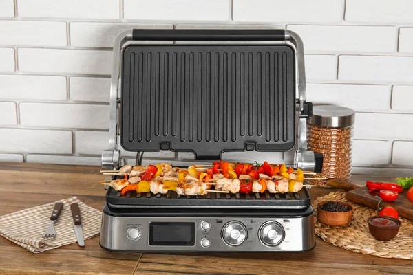 Modern electric grill with tasty chicken skewers and vegetables on table near light brick wall