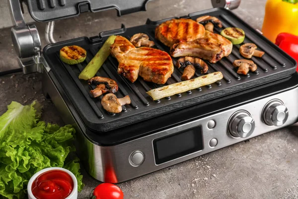 Modern electric grill with tasty steaks and vegetables on table