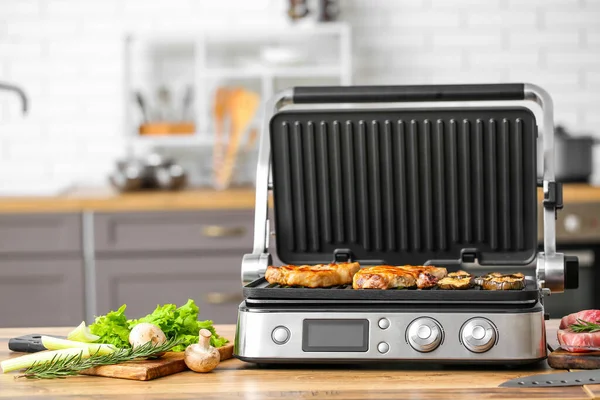 Modern electric grill with tasty steaks and vegetables on table in kitchen