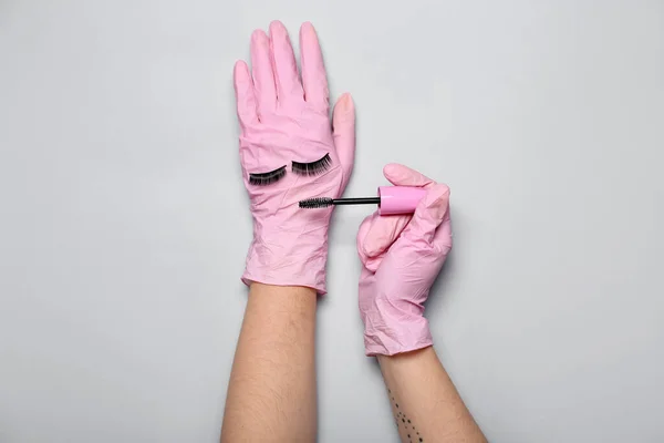 Makeup artist in pink gloves with mascara and fake eyelashes on grey background
