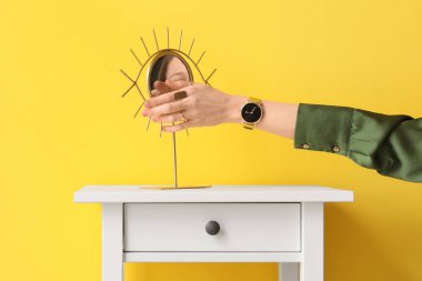 Woman with golden wristwatch and mirror on table near yellow wall