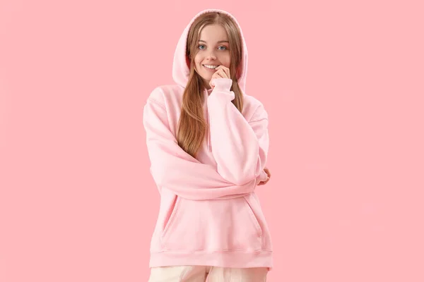 Young woman in hoodie biting nails on pink background