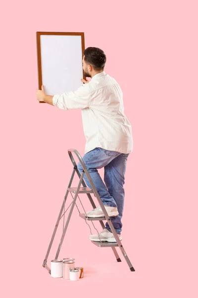 Young Man Ladder Hanging Frame Pink Background — Φωτογραφία Αρχείου