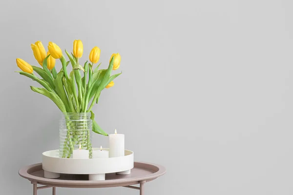 Burning candles and vase with tulip flowers on table near grey wall
