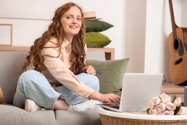 Güzel kızıl saçlı kadın evdeki kanepede dizüstü bilgisayar kullanıyor.