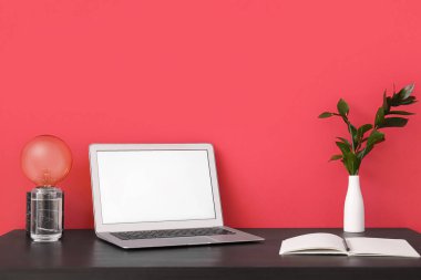 Kırmızı duvarın yanında dizüstü bilgisayarı, lambası ve defteri olan işyeri