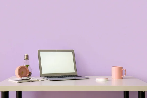 Workplace with laptop, cup and alarm clock near lilac wall