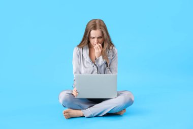 Young woman in pajamas with laptop biting nails on blue background