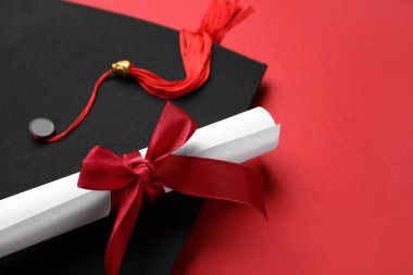 Kırmızı arka planda kurdeleli diploma ve mezuniyet şapkası.
