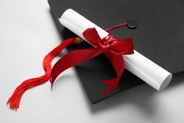 Kırmızı kurdeleli diploma ve beyaz arka planda mezuniyet şapkası.