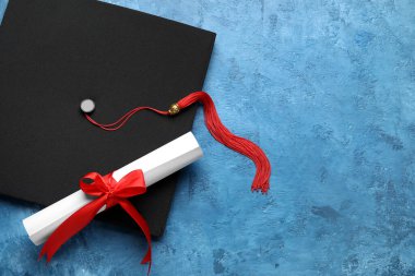 Diploması kırmızı kurdeleli ve mezuniyet şapkası mavi masada.