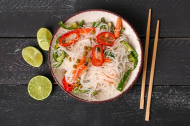 Bowl with tasty rice noodles, chopsticks, vegetables and lime on black wooden table clipart