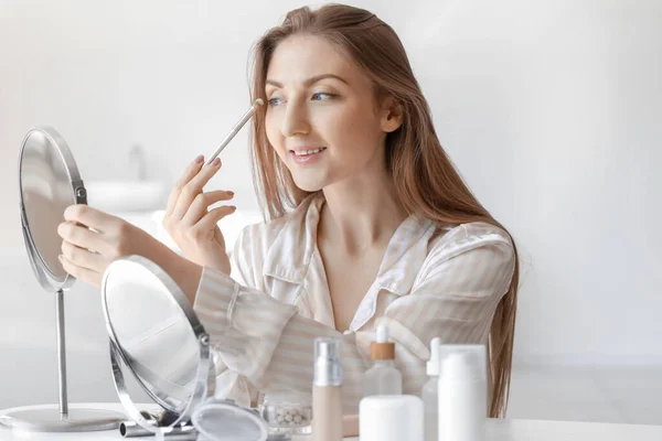 Young woman with mirror and brush applying eyeshadows at home