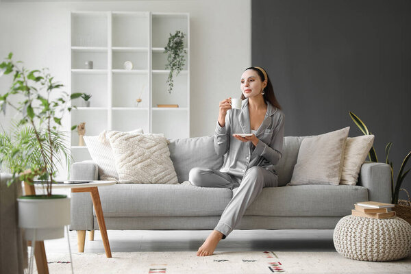 Young woman with sheet mask and cup of coffee sitting on sofa at home