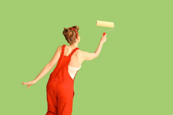 Young pin-up woman in red uniform with paint roller on green background, back view