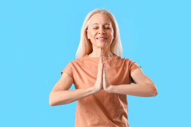 Olgun bir kadın açık mavi arka planda meditasyon yapıyor.