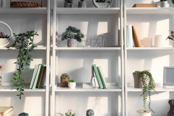 Shelving units with artificial plants, books and decor near light wall